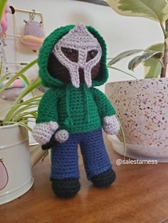 a crocheted doll with a green hoodie and blue pants stands on a table next to a potted plant