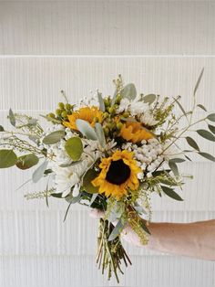 a person holding a bouquet of sunflowers and greenery in their hand, against a white wall