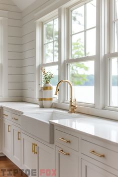 a kitchen with white cabinets and gold pulls on the faucet in front of two windows