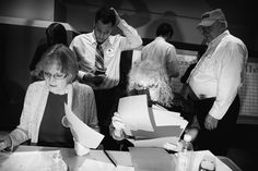 a group of people standing around a table with papers on it and one woman looking at her phone