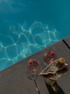 two wine glasses sitting on the edge of a swimming pool next to an empty bottle