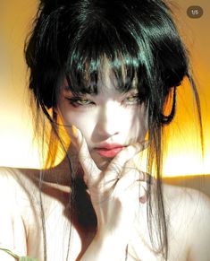 a woman with black hair and green eyes posing for the camera while holding her hand to her face