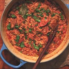 a blue pot filled with meat and vegetables on top of a wooden table next to a spoon