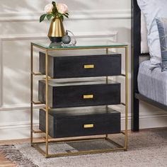 a black and gold side table with three drawers on it, next to a bed