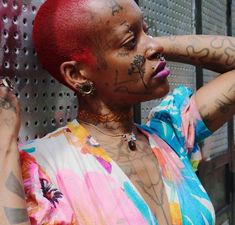 a woman with red hair and tattoos on her face, leaning against a metal wall