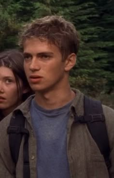 the young man and woman are standing together in front of some trees, one is looking at the camera