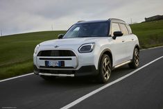 the front end of a white mini countryman driving on a road with grass and hills in the background