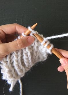 two hands are knitting together yarn on a black surface with a wooden stick in the foreground