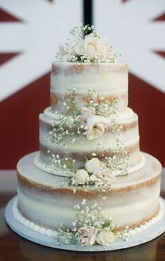 a three tiered wedding cake with white flowers on the top and bottom, sitting on a table in front of a british flag