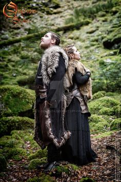 a man and woman dressed in medieval clothing standing on mossy ground with their arms around each other