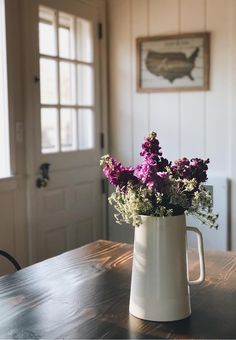 purple flowers in a white pitcher sitting on a wooden table next to a door and window