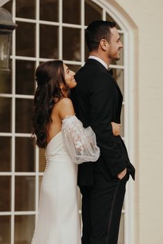 a bride and groom standing in front of a building with an arched window looking into each other's eyes