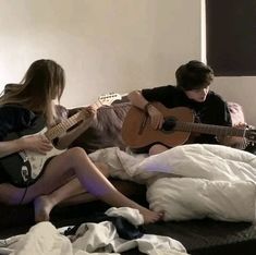 two people sitting on a bed playing guitar and another person laying down in the background