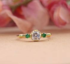 an engagement ring with three emeralds and a diamond in the center on a table next to flowers