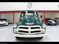 an old green truck parked in a garage next to other classic sports cars on display