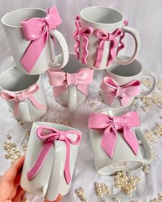 a hand holding a white and pink coffee mugs with bows on the handles, sitting next to other cups