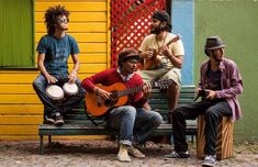 three men are sitting on a bench playing instruments and singing while another man plays the guitar