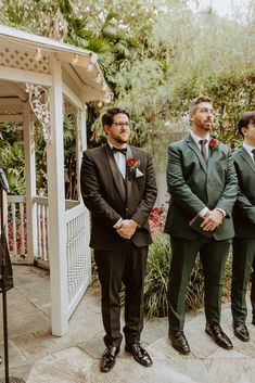 three men in suits standing next to each other near a gazebo with trees and bushes behind them