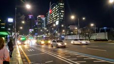 cars are driving down the street at night with tall buildings in the background and people walking on the sidewalk