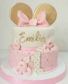 a pink and white cake decorated with minnie mouse ears, gold nameplates and the number one on top