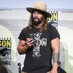 a man with long hair wearing a hat standing in front of a table holding a cell phone