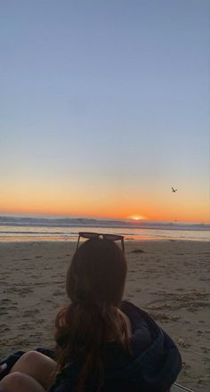 a woman sitting on top of a sandy beach next to the ocean at sun set