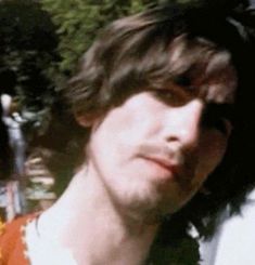 a man with long hair wearing an orange and white shirt standing in front of trees