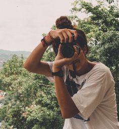 a man holding a camera up to his face with trees in the back ground behind him