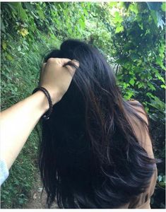 a woman with long black hair standing in front of some trees and bushes holding her hand up to her head