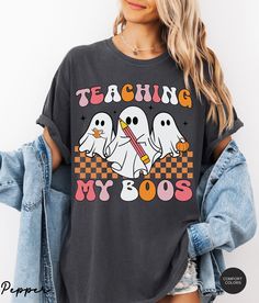 Get ready for a spooky season in style with this Comfort Colors® Teacher Halloween Shirt. This Retro Ghost Teacher Shirt, featuring the fun "Teaching My Boos" design, is the perfect Halloween Teacher Shirt for any classroom, making it a must-have Spooky Teacher T-shirt. Comfort Colors® Teacher Halloween Shirt, Halloween Teacher Shirt, Teaching My Boos Shirt, Spooky Teacher T-shirt, Retro Ghost Teacher Shirt About Comfort Colors C1717 The Models in the pictures are wearing 1 or 2 sizes up, Please Casual Halloween Graphic Tops, Casual Graphic Tops For Halloween, Casual Halloween Shirt With Graphic Design, Funny Graphic Print Shirt For Fall, Halloween Graphic Tee Shirt With Graphic Design, Halloween Graphic Tee With Graphic Design, Fall Graphic Tee Shirt With Character Print, Fall Graphic Tee With Character Print, Short Sleeve Halloween Graphic Shirt