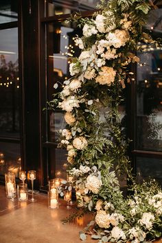 an arrangement of white flowers and greenery with candles in front of a large window