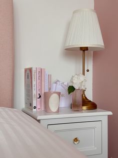 a white nightstand topped with books and a lamp next to a pink headboard in a bedroom