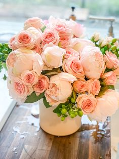 a white vase filled with pink roses on top of a wooden table next to a window