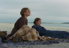 two people are sitting on the beach under blankets and looking at something in the distance