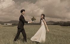 a man and woman are walking through the grass holding hands with each other in front of an overcast sky