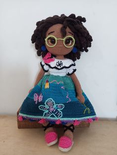 a crocheted doll with glasses sitting on a wooden table next to a white wall