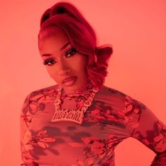 a woman with red hair wearing a floral shirt and necklace, standing in front of a pink background