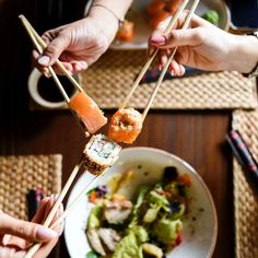 two hands holding chopsticks with sushi and vegetables on the plate in front of them