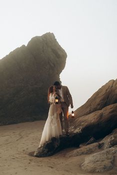 a bride and groom standing on the beach holding candles in front of a rock formation