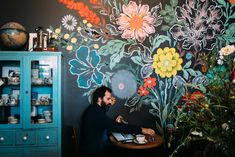 a man sitting at a table in front of a wall with flowers painted on it