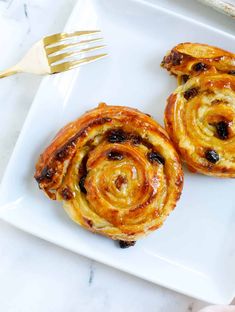 two cinnamon rolls on a white plate with a gold fork and knife next to it