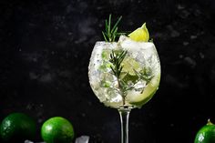 a gin cocktail garnished with rosemary and limes on a black table next to lemons