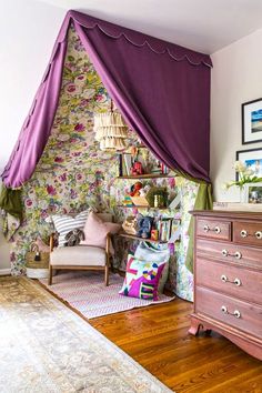 a bedroom with purple curtains and floral wallpaper on the walls, along with an old chest of drawers