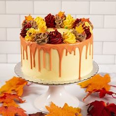 a cake with orange icing and flowers on it next to fall leaves in front of a white brick wall