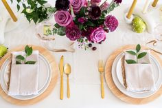 the table is set with white plates, silverware and purple flowers in vases