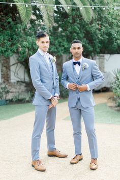 two men in suits standing next to each other on a gravel ground with palm trees behind them