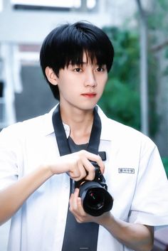 a man holding a camera and wearing a neck tie in front of a building with trees