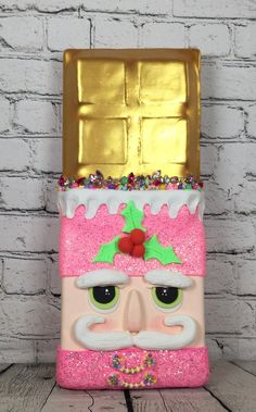 a pink and gold box with a santa clause on it's head, sitting next to a white brick wall