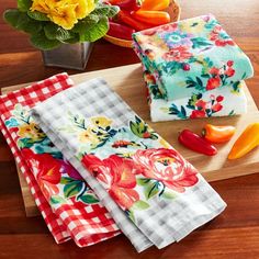 four colorful napkins on a cutting board with peppers and flowers in the background,