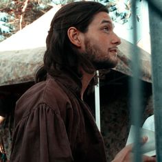 a man with long hair standing in front of a fence and looking off into the distance